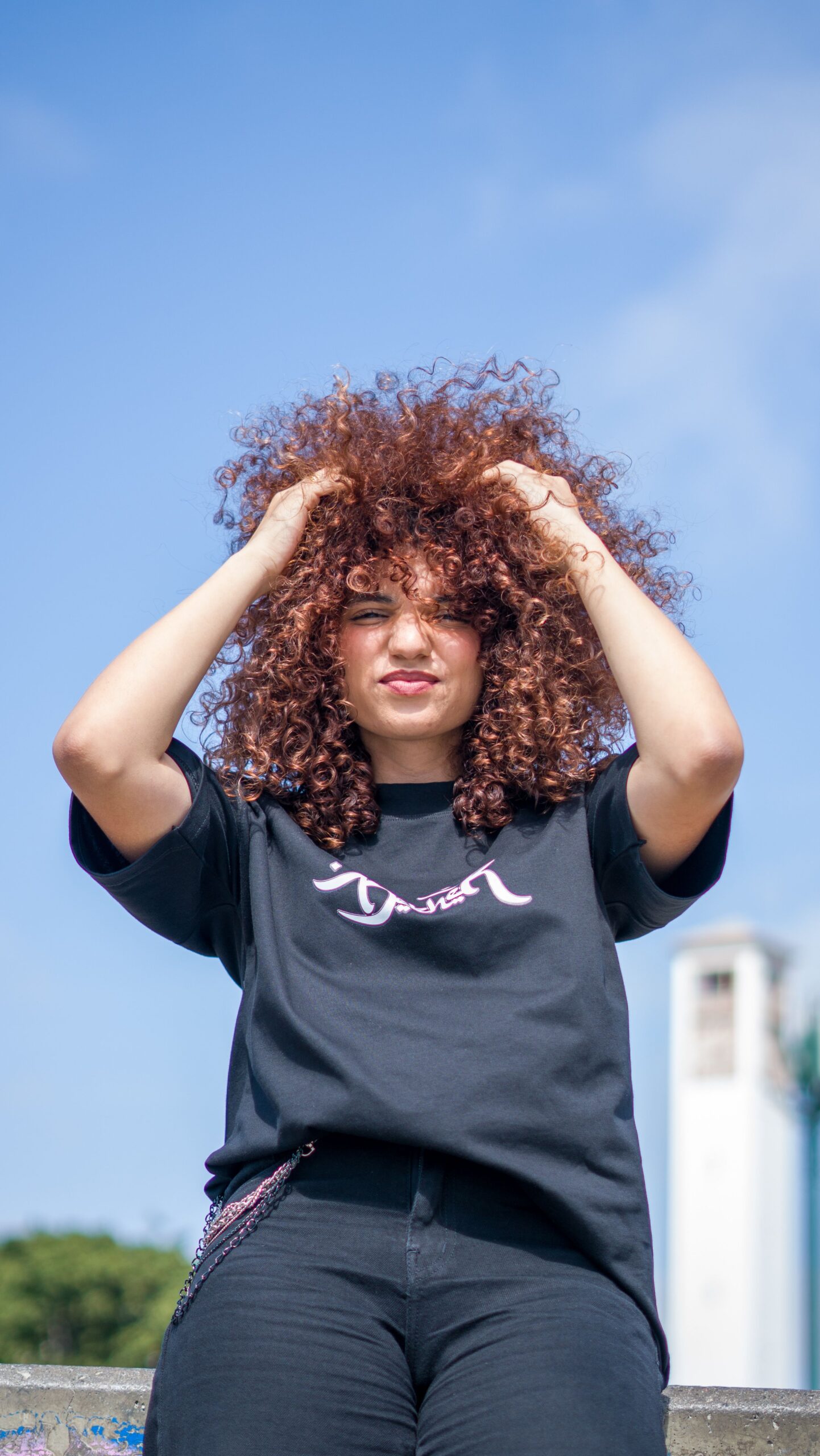 Confident young woman with curly hair posing outdoors with a blue sky backdrop, embracing natural beauty.